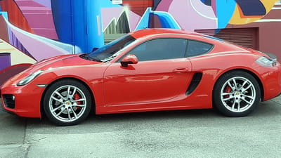 Porsche Cayman mit Original-Felgen von OR24.de Porsche Cayman in Rot vor einer bunten Graffiti-Wand, Seitenansicht mit silbernen Original-Felgen im 5-Doppelspeichen-Design