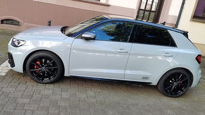 Audi A1 auf Original-Felgen Audi A1 in Grau mit schwarzem Dach, schräg von vorne am Gehweg, auf originalen 10-Speichen-Felgen in Schwarz